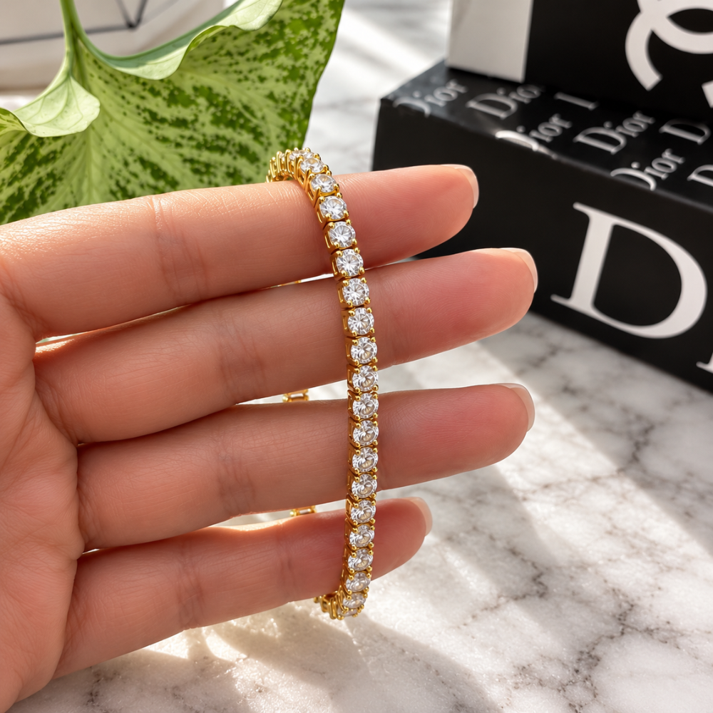 Close-up of MARLA gold-tone moissanite tennis bracelet held between fingers, showcasing brilliant stones and elegant gold settings.
