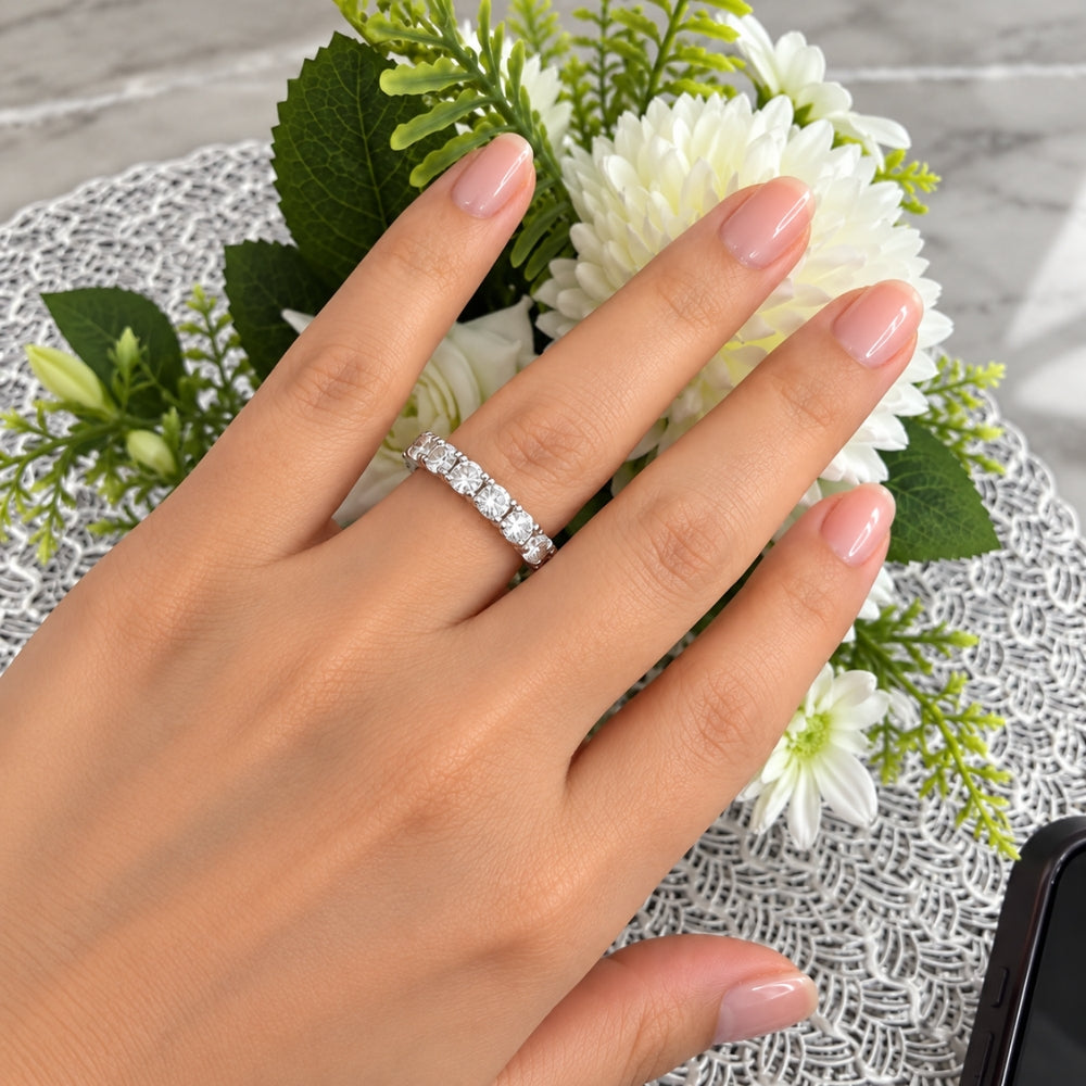 Marla Jewellery Moissanite Eternity Ring worn on a model's hand with glossy pink nails, surrounded by elegant white flowers and green foliage.
