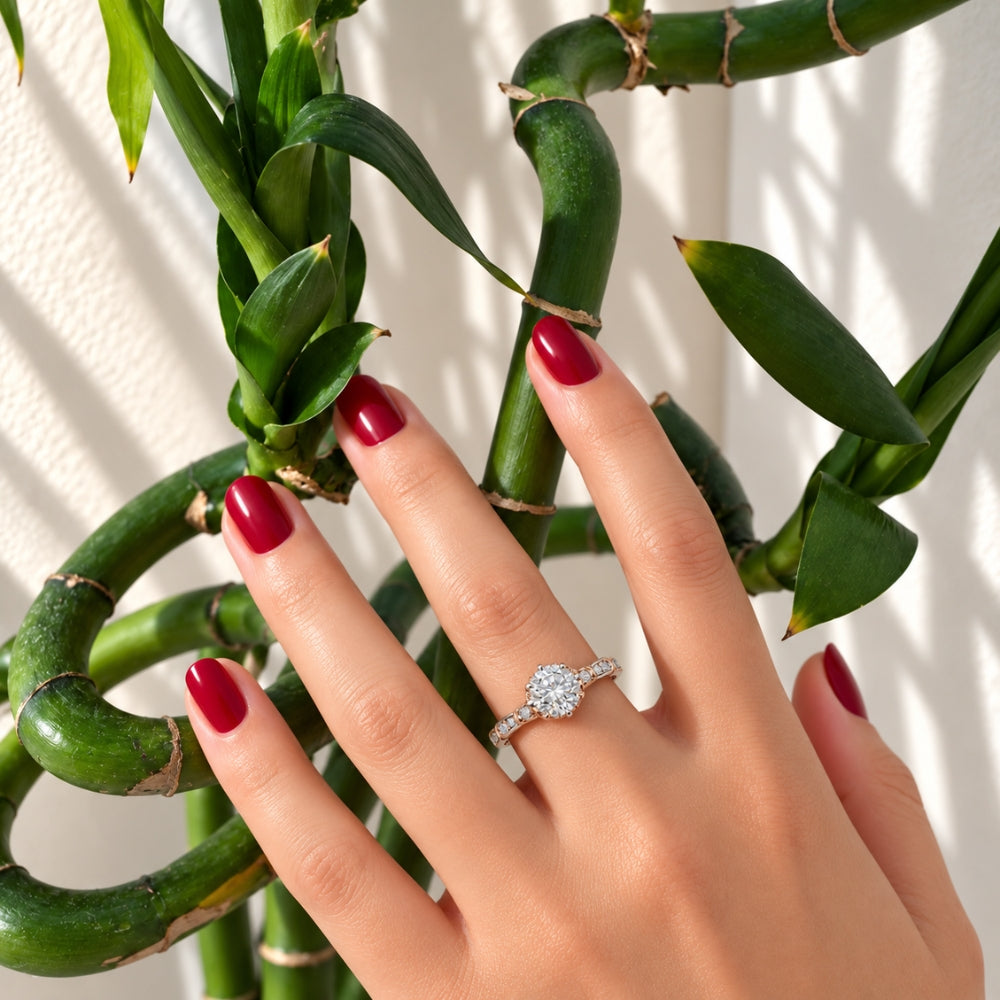 Marla Rose Light Ring with sparkling 1ct round moissanite stone in 925 sterling silver, worn on a woman's hand with burgundy nails against green bamboo.